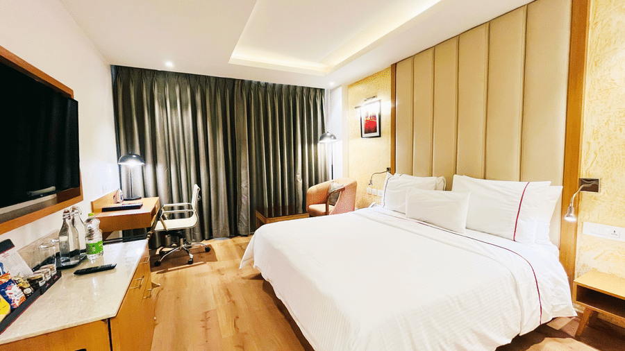 A hotel bedroom with a large bed, a work desk with a chair, dark grey curtains, and a television mounted on the wall.