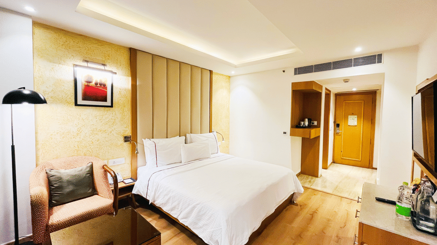 A warm hotel room interior showing a white bed, an armchair in the corner, and a hallway leading to the entrance door.