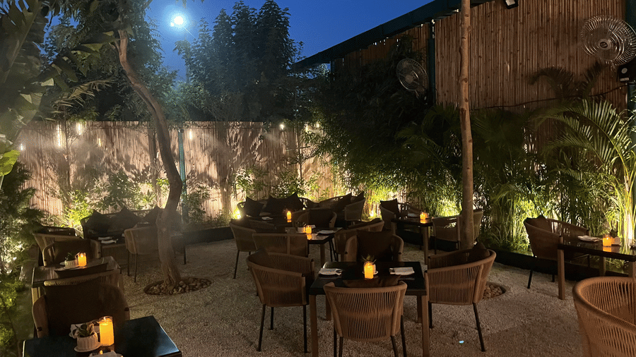 Atmospheric outdoor garden restaurant at night featuring wooden tables, woven chairs, ambient candle lighting, and lush green trees.