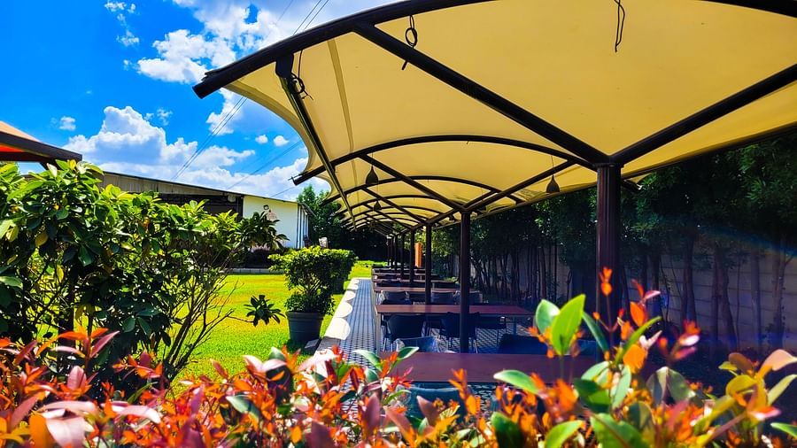 Covered outdoor dining area with tables lined under a beige canopy amidst lush greenery. | Vistar Resort, Bengaluru