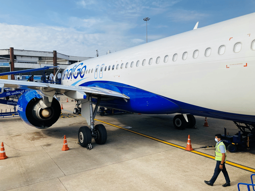 Airplane parked on airport runway with ground staff, cones, and airport equipment visible around the aircraft.