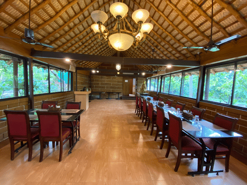 Formal dining setup at Ibex River Resort, Pollachi showcasing wooden tables and chairs, elegant chandeliers, and large windows providing natural light and scenic forested views for guests.