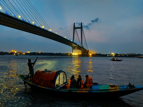 A boat with people on it travelling on a water body in front of the Howrah Bridge in Kolkata during twilight hour.