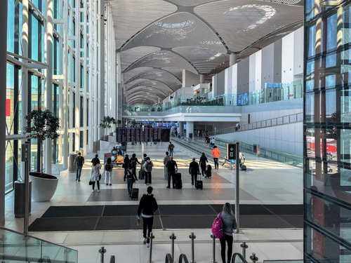 Airport terminal interior with people walking, glass walls, overhead structure, and seating areas along the passage.