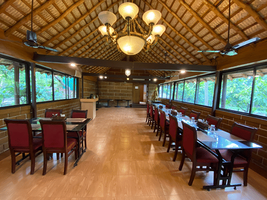 Formal dining setup at Ibex River Resort, Pollachi featuring wooden tables and chairs, elegant chandeliers, and large windows providing natural light and scenic forested views for guests.