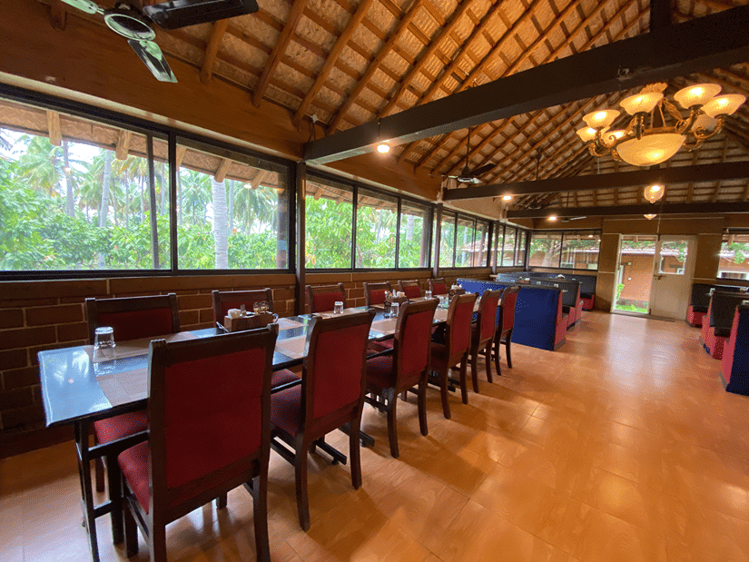 Elegant dining area at Ibex River Resort, Pollachi with neatly arranged wooden tables and chairs, large windows offering views of the surrounding greenery, and warm ambient lighting.