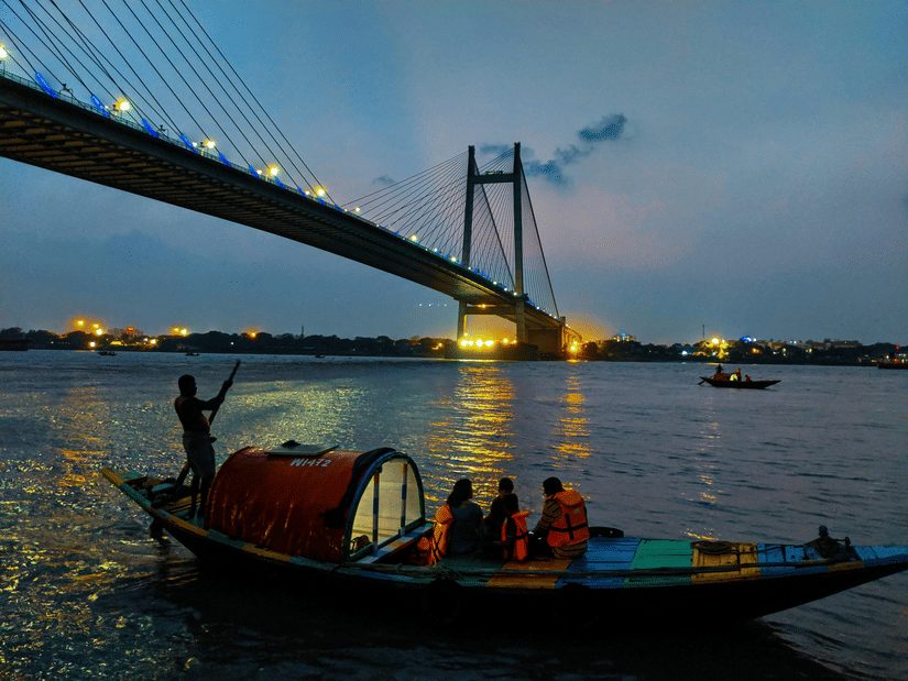 A boat with people on it travelling on a water body in front of the Howrah Bridge in Kolkata during twilight hour.