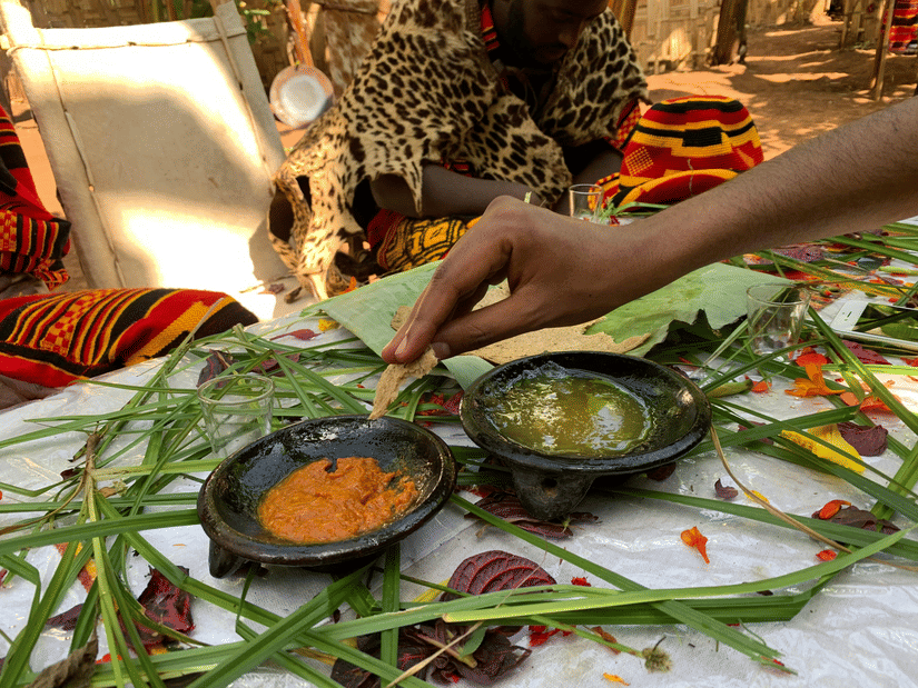 A person using a injera, an Ethiopian traditional food, to dip into one of the two bowls of sides with two people sitting in front of the table