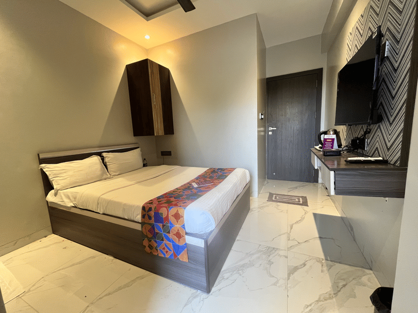 A compact modern bedroom features a neatly made bed, tiled flooring, warm lighting, a wall-mounted television and a functional desk area at VITS Select Kharadi, Pune.