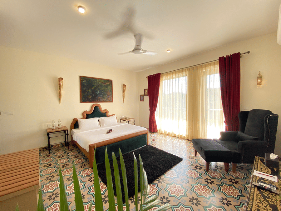 A bedroom with patterned floor tiles, a bed with a headboard, and an armchair. A ceiling fan, two wall sconces, a painting, two bedside tables, and large glass windows with curtains - Karma Lakelands