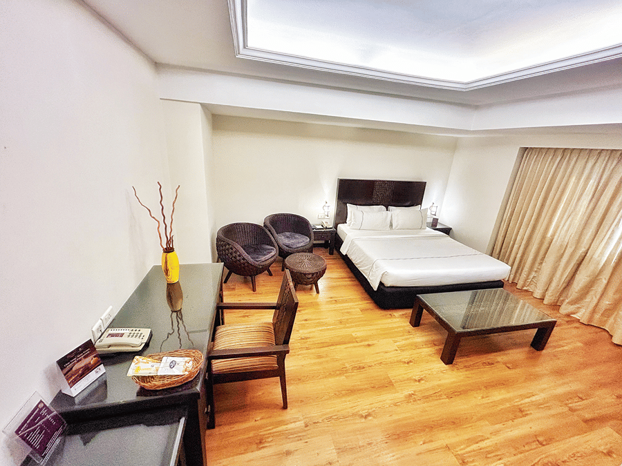 Residency Suite at The Residency Karur shows a double bed, a glass coffee table, two round chairs, and a desk with a wooden chair on a wooden floor.