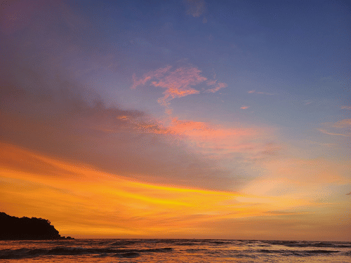 A view from the shoreline of Cavelossim Beach after sunset with different coloured hues in the sky and a hill on the left hand side.