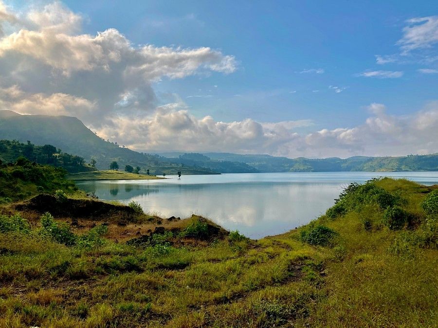 Scenic view of a calm lake reflecting blue skies and clouds, surrounded by green hills and sparse trees under a sunny sky.