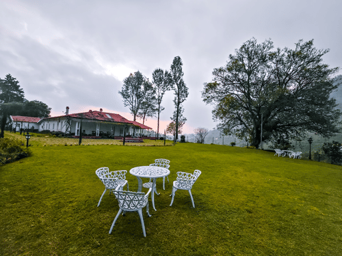  A serene outdoor setting featuring a white table and four chairs on a lush green lawn, with a colonial-style building and trees under a cloudy sky in the background.