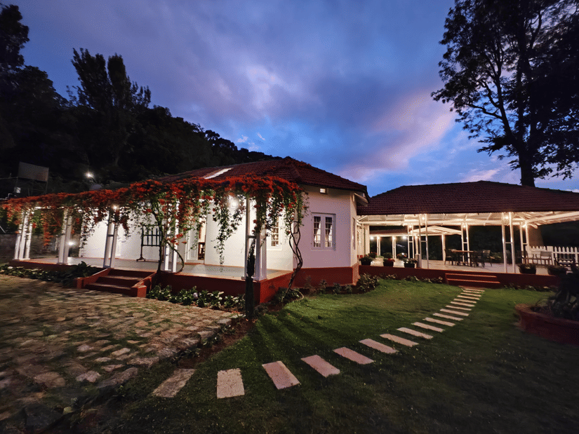 A long, single-story building with a red roof and an illuminated veranda covered in vines, with stepping stones on a path leading across a lawn at dusk.