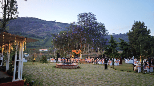 Wide shot of an outdoor wedding venue with guests seated around a central stage under a large tree with purple flowers in Coonoor - Ibex Resorts, Coonoor (Leewood).
