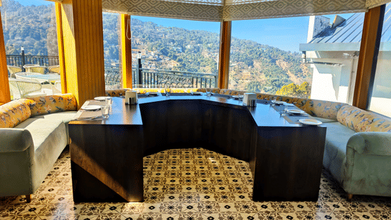 Curved booth seating with floral cushions around a dark table, with panoramic windows overlooking Kasauli hillside