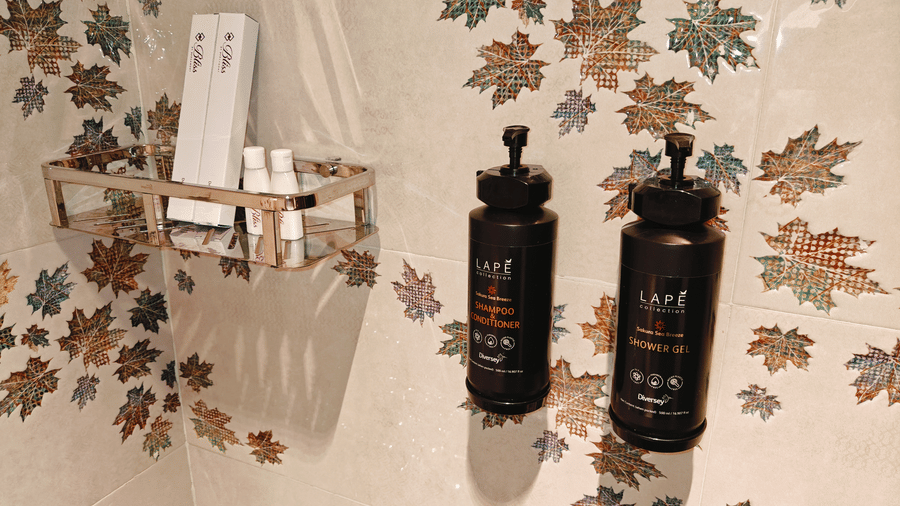 Black dispensers and white toiletry boxes kept on a glass shelf against a leaf-patterned wall at Bliss Casa Mila By Bergamont Hotels, Kodaikanal.