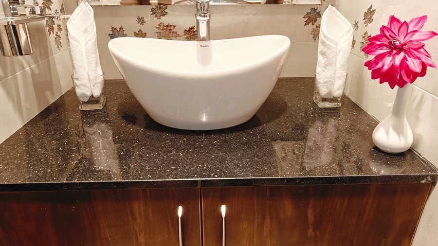 A white oval washbasin sits on a dark granite counter with 2 rolled white towels and a pink flower at Bliss Casa Mila By Bergamont Hotels, Kodaikanal.