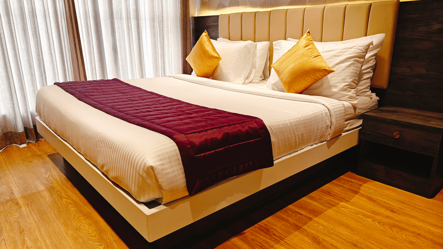 A bed with 2 white pillows, 2 yellow cushions, and a maroon runner is shown from a side angle at Bliss Casa Mila By Bergamont Hotels, Kodaikanal.