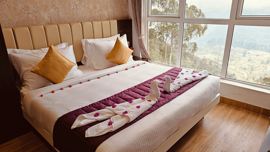 A bed features white towels folded into a pair of swans and scattered red petals in front of a window at Bliss Casa Mila By Bergamont Hotels, Kodaikanal.