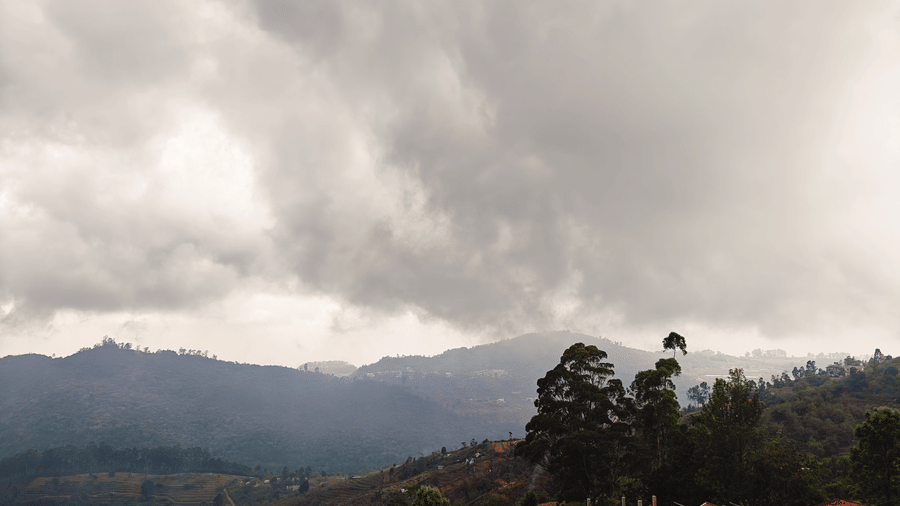 View of grey clouds hang over a dark green forest on a hilly landscape during the day from Bliss Casa Mila By Bergamont Hotels, Kodaikanal.