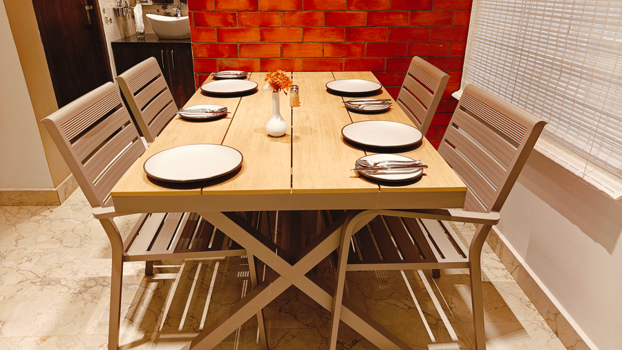A light wood dining table set for 6 people with white plates and cutlery is placed against a red brick wall at Bliss Casa Mila By Bergamont Hotels, Kodaikanal.