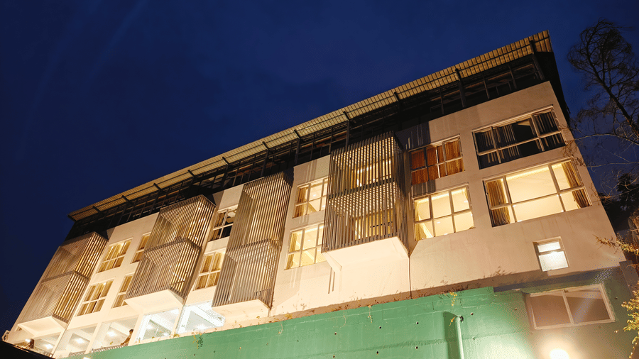 A 4-storey white hotel building with lit windows and a green base viewed at night at Bliss Casa Mila By Bergamont Hotels, Kodaikanal.