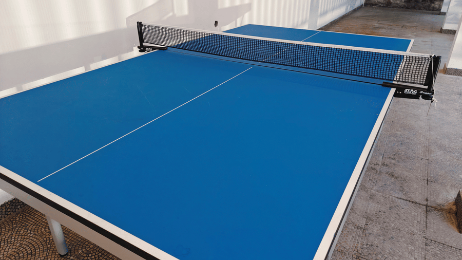 A blue table tennis table with a black net stands on a grey floor indoors at Bliss Casa Mila By Bergamont Hotels, Kodaikanal.