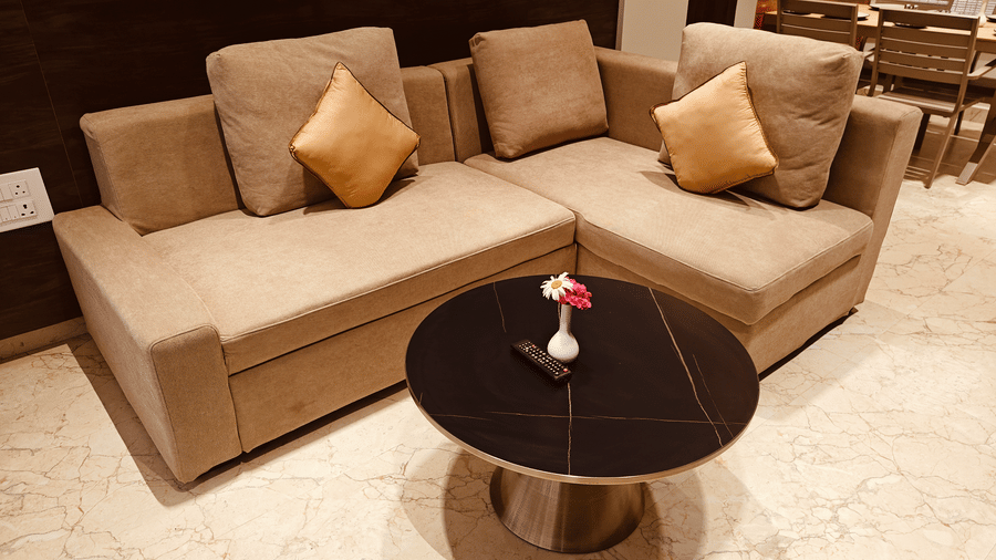 A beige L-shaped sofa with yellow cushions faces a round black coffee table on a tiled floor at Bliss Casa Mila By Bergamont Hotels, Kodaikanal.