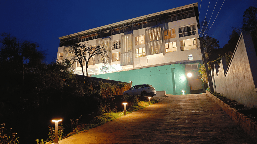 A paved driveway lit by 2 small lamps leads towards a large white building at night at Bliss Casa Mila By Bergamont Hotels, Kodaikanal.