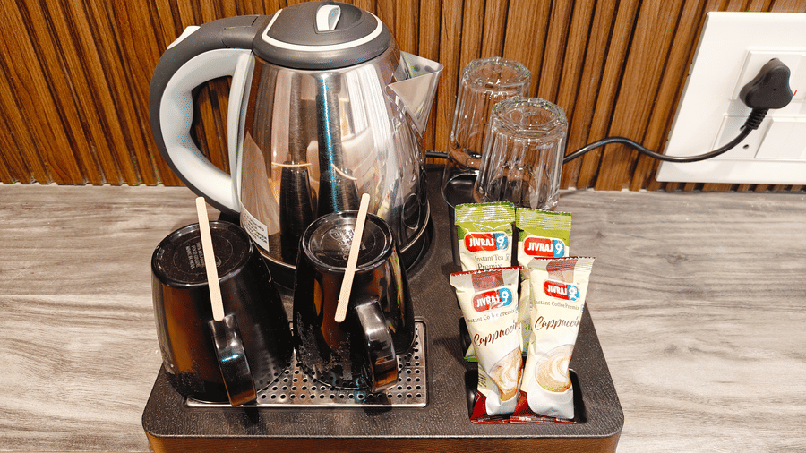 A silver electric kettle, 2 black mugs, 2 glasses, and 4 coffee sachets sit on a black tray at Bliss Casa Mila By Bergamont Hotels, Kodaikanal.