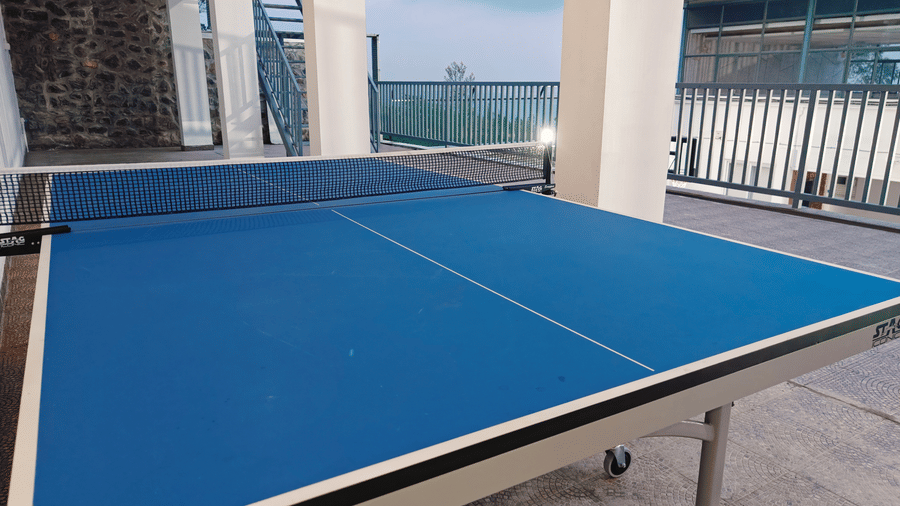 A blue indoor table tennis table with a net viewed from 1 end on a tiled floor at Bliss Casa Mila By Bergamont Hotels, Kodaikanal.