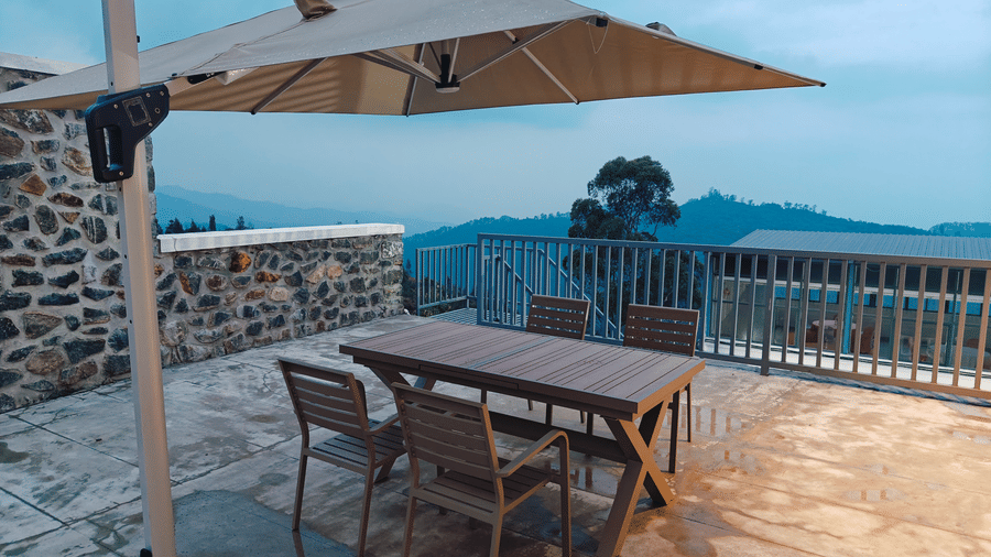 A wooden table and 4 chairs under a large beige umbrella on a stone terrace at Bliss Casa Mila By Bergamont Hotels, Kodaikanal.