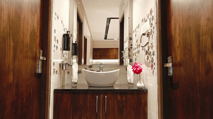 A view through wooden doors shows a white washbasin and a pink flower in a bathroom with patterned tiles at Bliss Casa Mila By Bergamont Hotels, Kodaikanal.