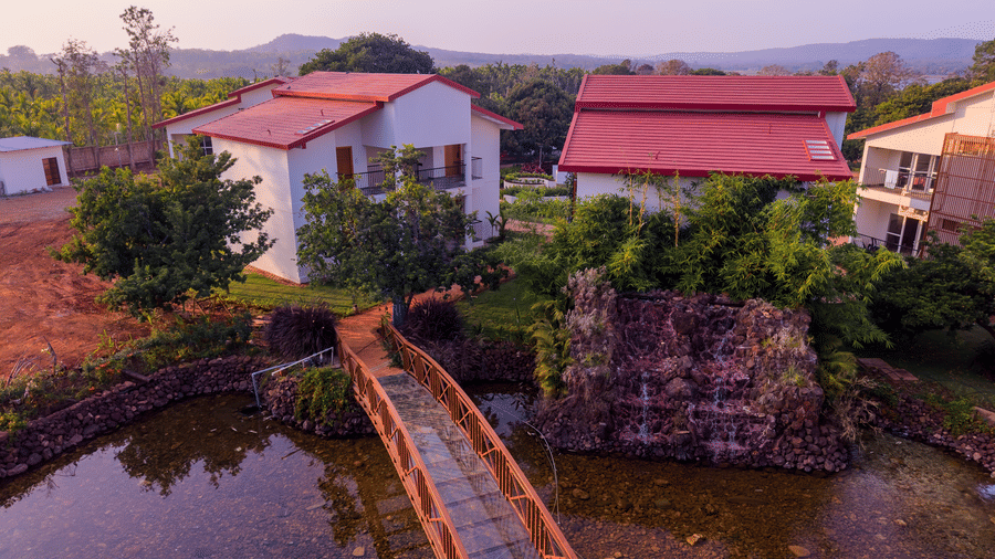 An elevated view of a brick walkway bridge built across a small pond leading toward red-roofed resort villas | Lords Sahyadri Resort, Shivamogga