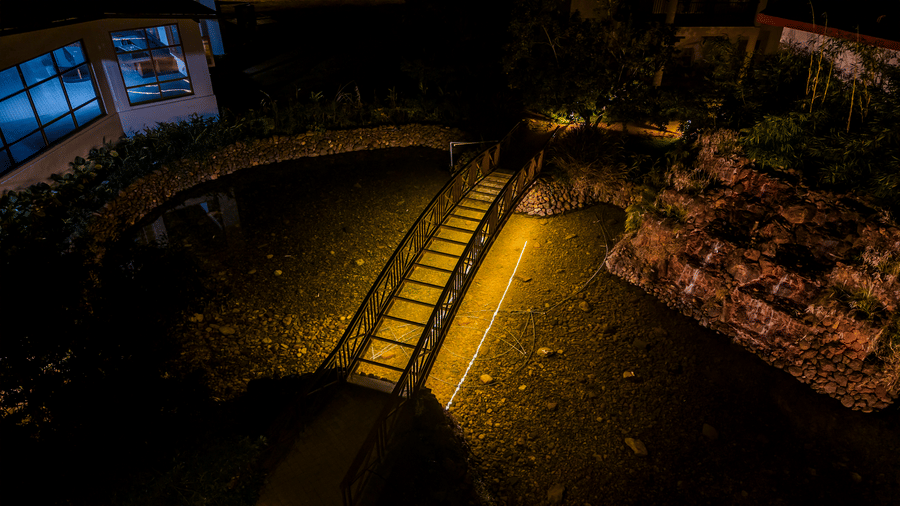 A night view of a small wooden bridge over water, illuminated by a spotlight from above | Lords Sahyadri Resort, Shivamogga
