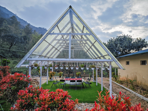 A white-framed, transparent-roofed gazebo or greenhouse structure with a green lawn inside set for dining, surrounded by red and green plants, with misty mountains in the background under a cloudy sky.