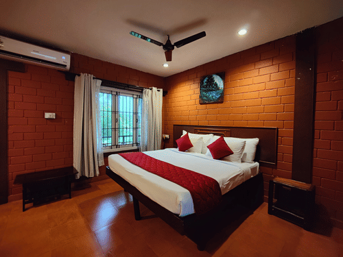 A double bed with open brick style wall behind the bed and a ceiling fan hanging from above inside the Executive Deluxe Rooms near Aliyar Dam at Ibex River Resort, Pollachi.