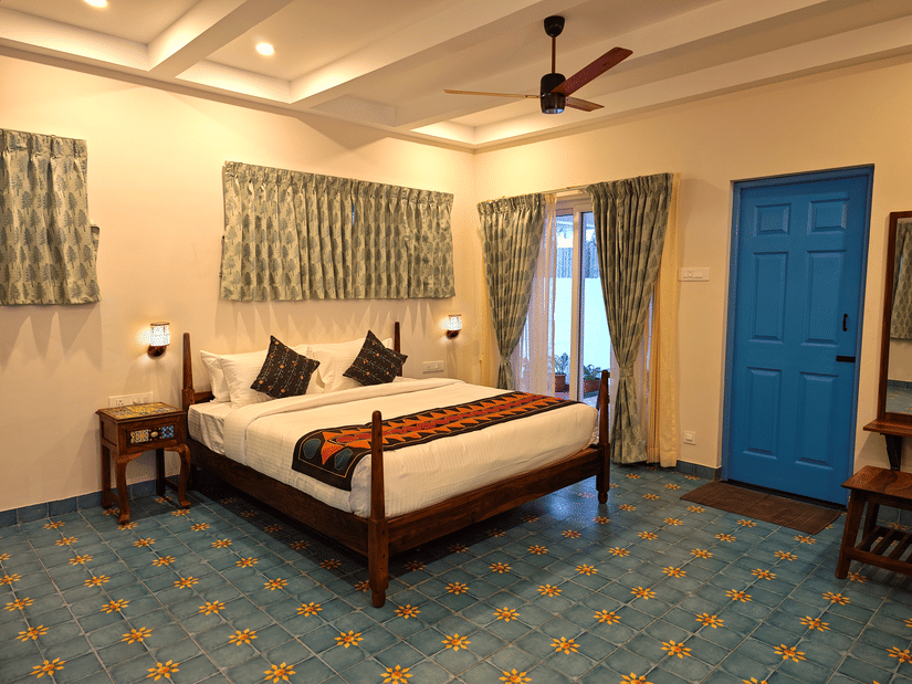  A well-appointed bedroom with a large bed, wooden frame, and colorful throw, complemented by a blue tiled floor, a blue door, and patterned curtains.