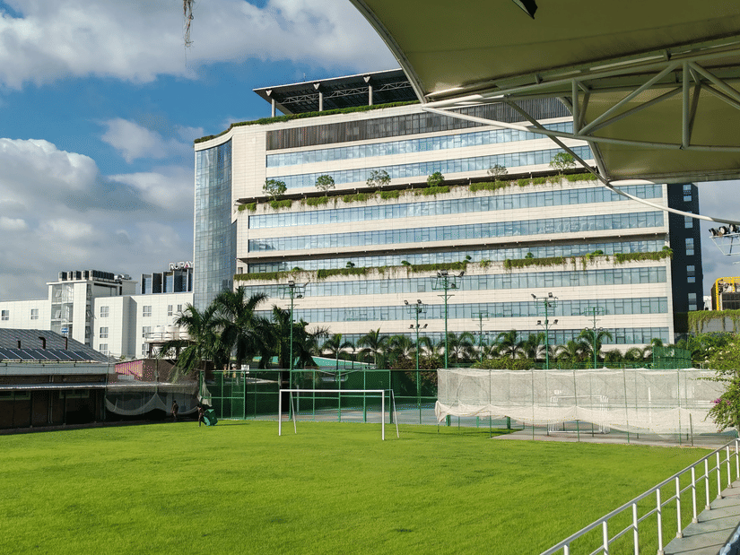 A large office building sits next to an open green sports field with a soccer goal, under a blue sky with white clouds.