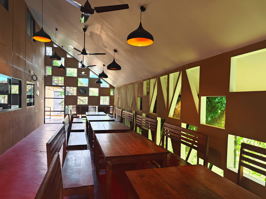 Interior of a rustic dining hall with long wooden tables, warm pendant lighting, and a unique geometric window wall - Ibex Resorts, Malampuzha (Kava Eco Camp and Caravan Park)