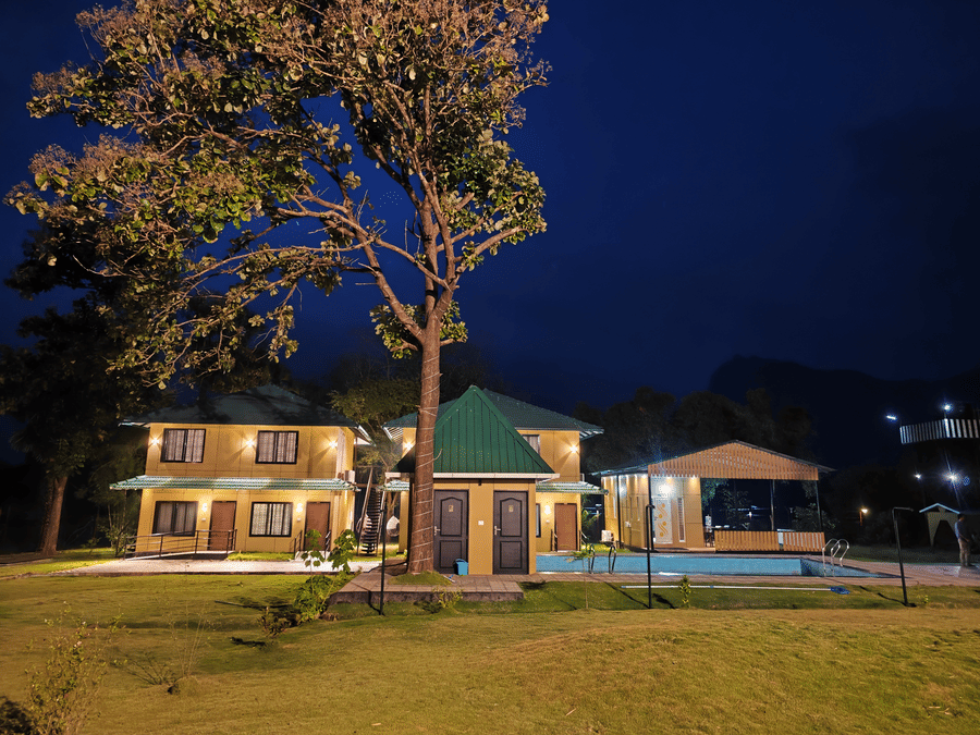 A warm night shot of the resort buildings illuminated against a dark blue sky with a tall tree in the foreground - Ibex Resorts, Malampuzha (Kava Eco Camp and Caravan Park)