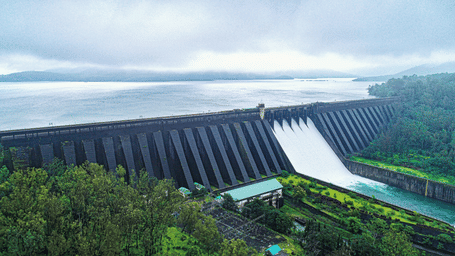 A dam with multiple gates releasing water into a channel, surrounded by trees and greenery, with cloudy sky above and water reservoir behind.
