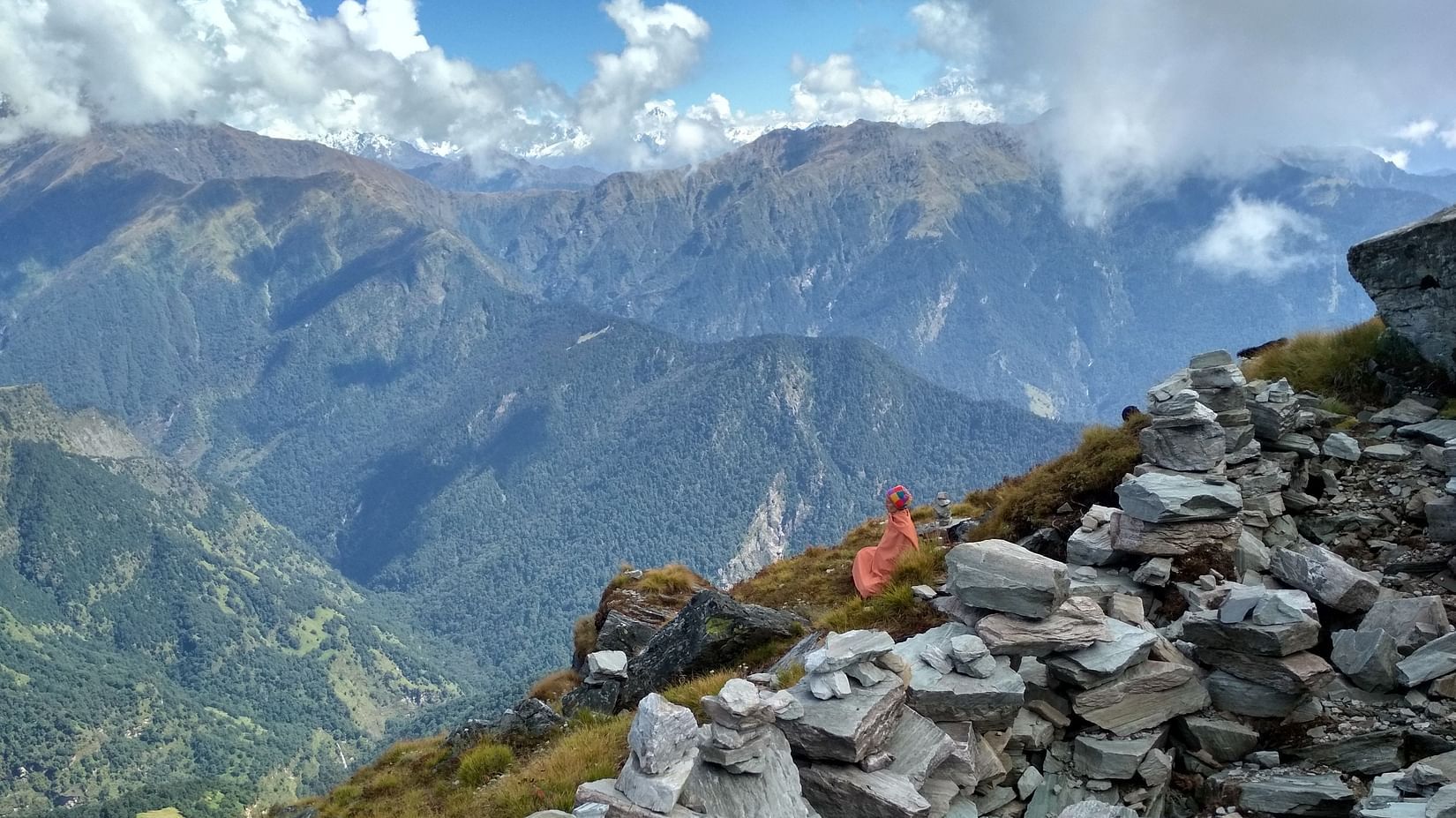 Several colourful tents are pitched on a rugged, rocky mountain terrain, with majestic snow-capped peaks visible in the distance under a partly cloudy sky, indicating a high-altitude camping expedition