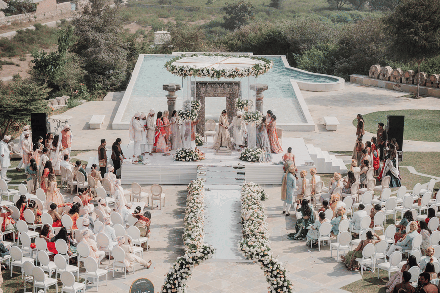 A wedding ceremony at the outdoor featuring as aisle decorated with flowers on either side, guests seated on either side and a centre stage where the rituals are taking place  with bride, broom, and their close family members.