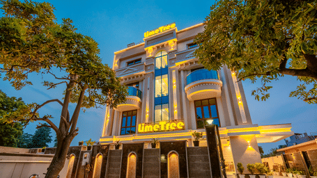 Ornate multiple storey building exterior of Lime Tree Hotels & Service Apartment Private Limited illuminated at dusk with surrounding trees.