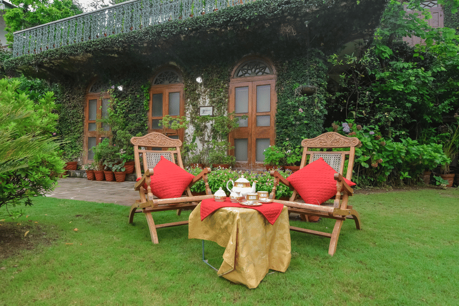 An outdoor seating in the gardens of resort featuring to wodden chairs and a small wooden table with a tea pot set resting on it.