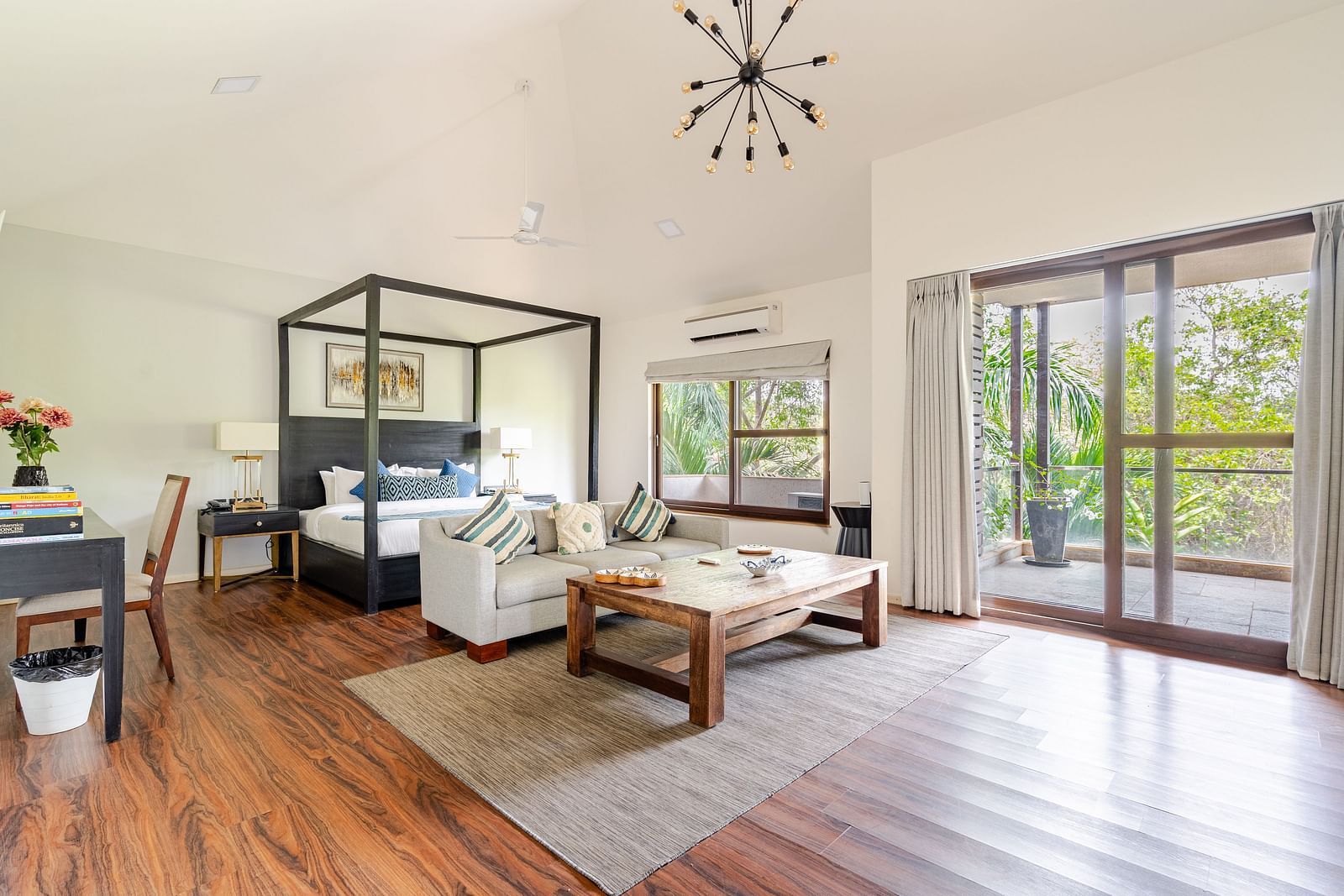 Spacious, airy bedroom with high ceilings and warm wood flooring, featuring a four-poster bed with patterned pillows, a modern gray sectional sofa, a rustic wooden coffee table atop a neutral rug, a writing desk with books and flowers, and large glass door