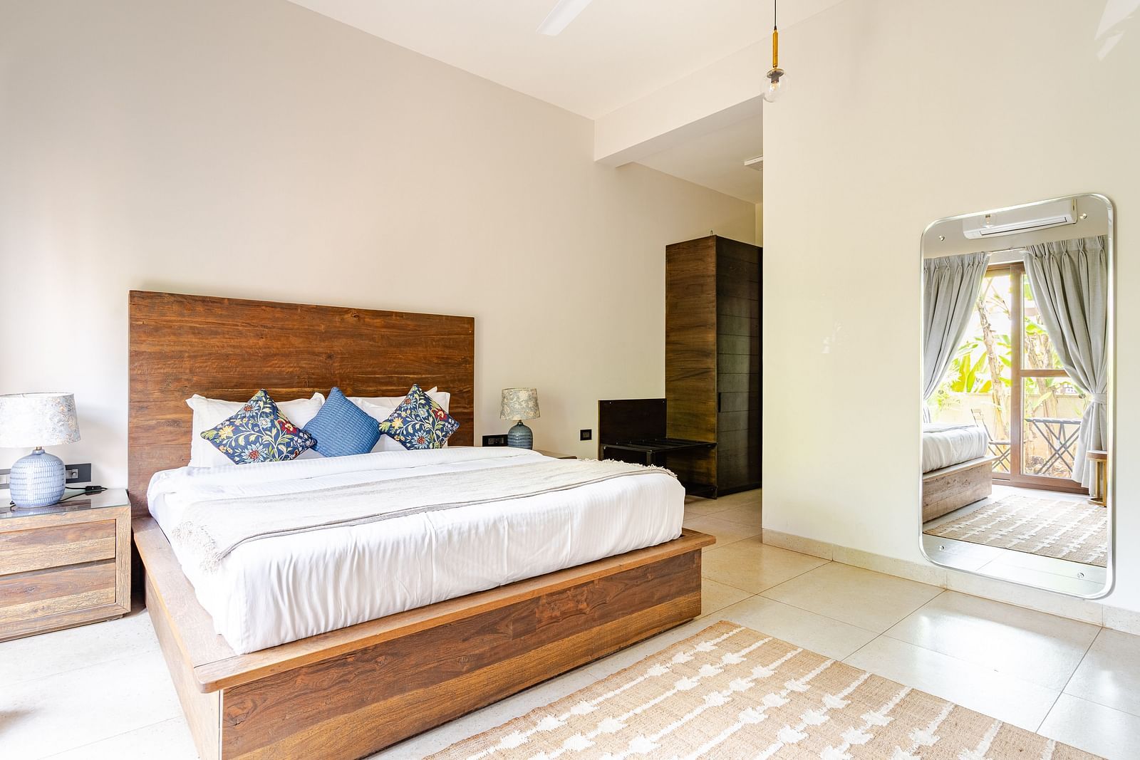 Bright and spacious bedroom featuring a wooden bed with patterned throw pillows, matching side tables, a full-length mirror reflecting natural light from a balcony door, and light-coloured tiled flooring - Lindsay Manor